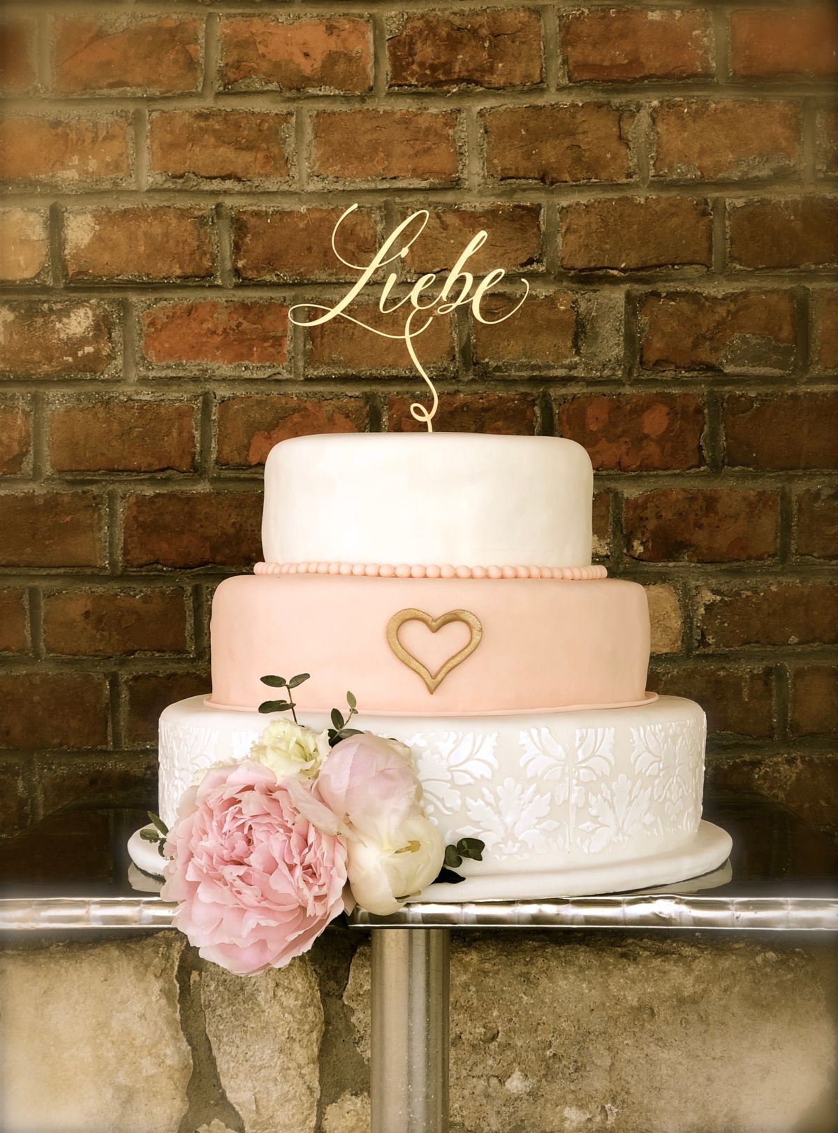 Dreistöckige Hochzeitstorte mit weißer Oberetage, rosa Mittelebene und Goldherz, Pfingstrose auf Ziegelwand