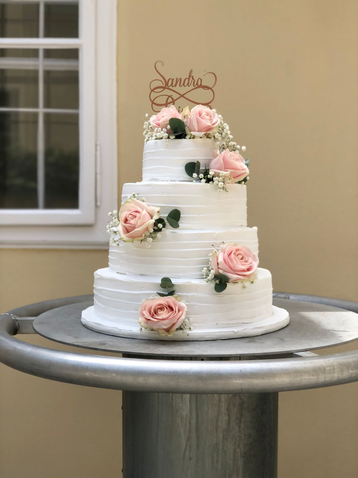 Four-tier white wedding cake with a gold name topper and cascading pink roses