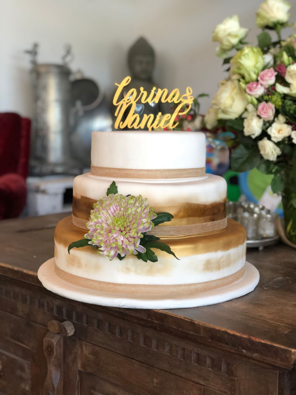 Three-tier white wedding cake with gold name topper and pale pink dahlia flower