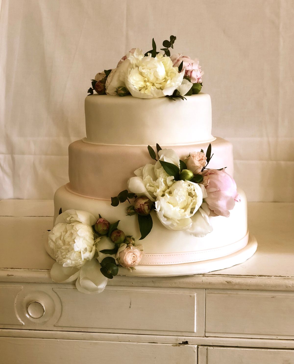 Three-tier wedding cake with white and blush fondant, fresh peonies and eucalyptus leaves