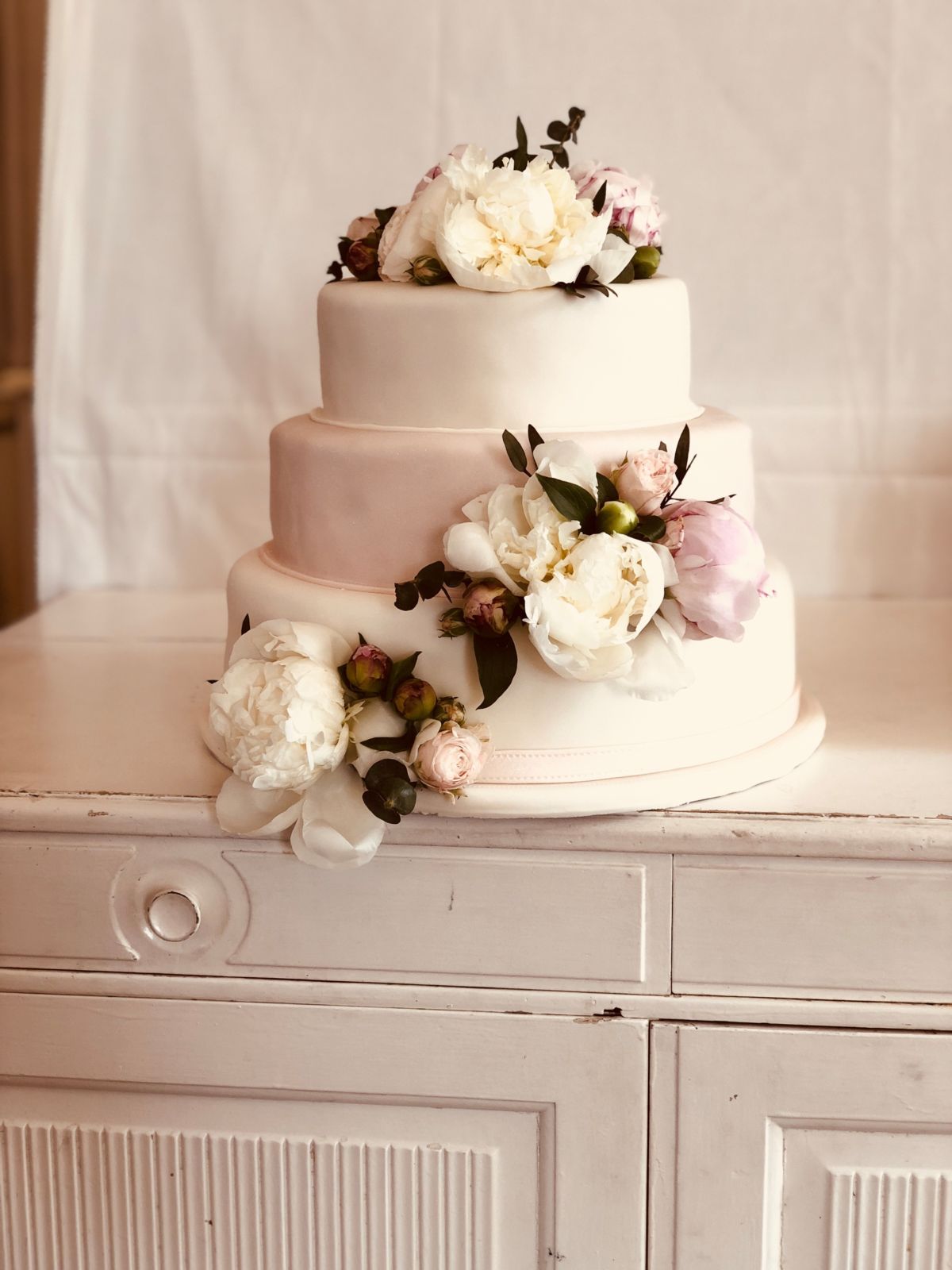 Three-tier wedding cake with blush middle tier, white peonies and eucalyptus on wooden table