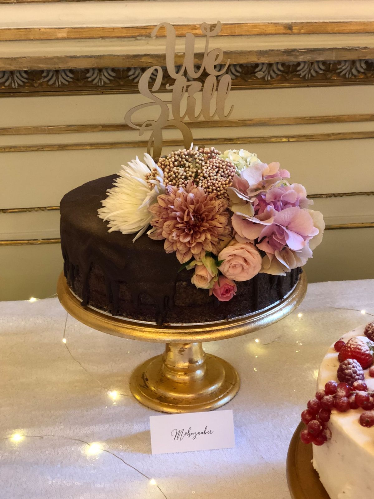 Single-tier dark chocolate cake on gold stand with ‘oh girl’ topper and mauve flower arrangement