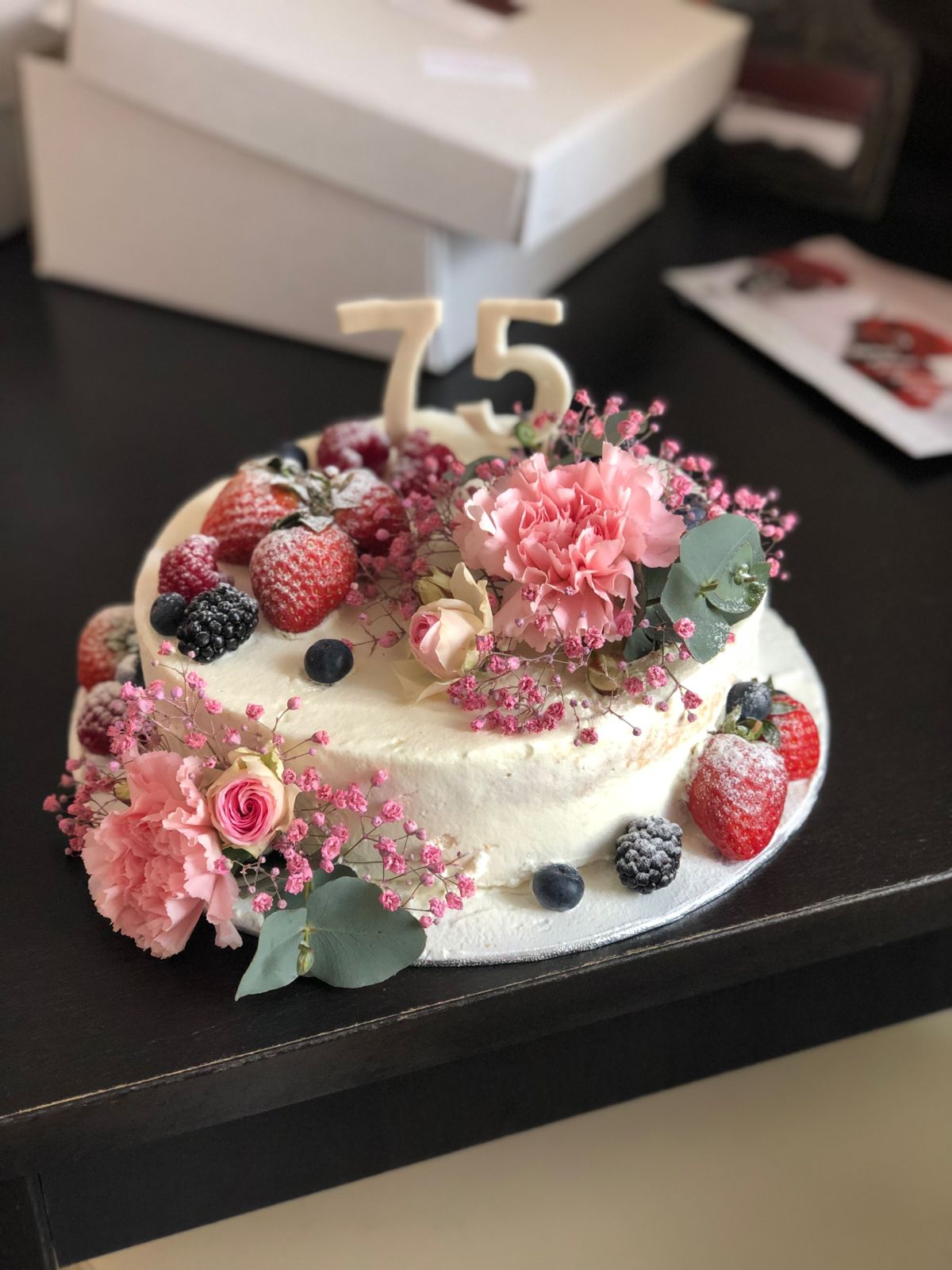 White frosted cake topped with fresh berries, pink flowers, and gold number 75