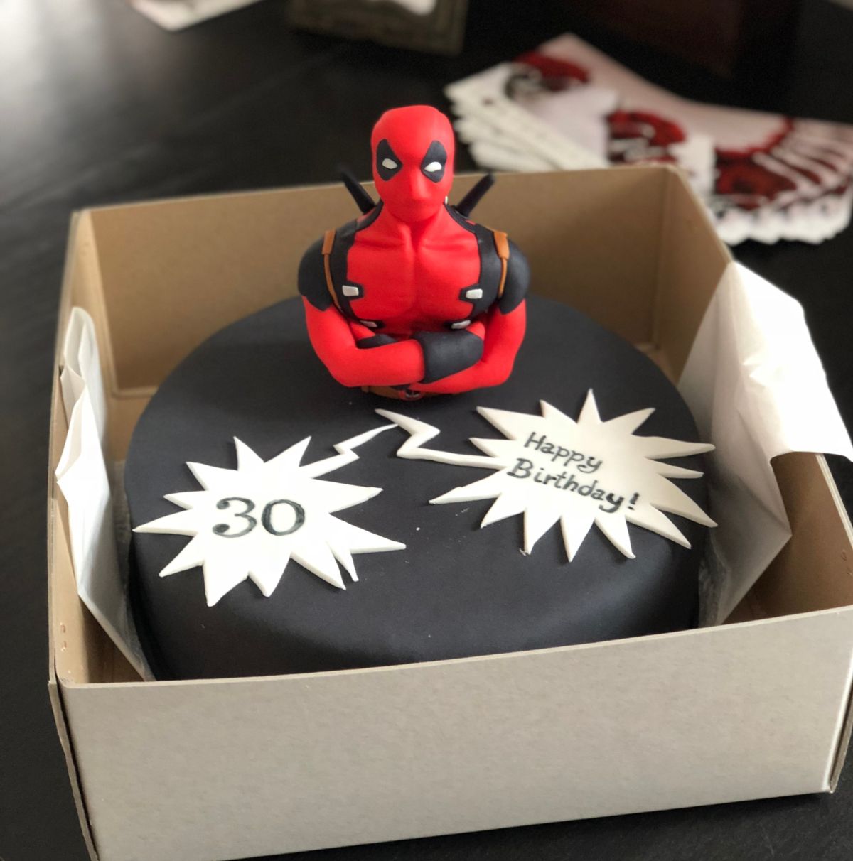 Adult birthday cake with red and black Deadpool fondant figure on dark base and comic-book burst designs