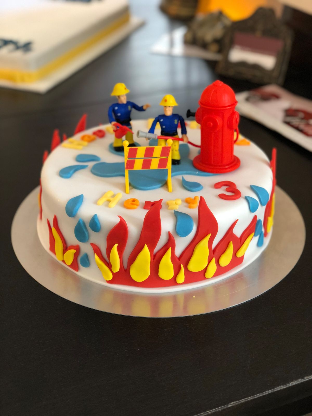 Firefighter-themed children’s birthday cake with miniature firefighter figures, red hydrant model, and flame decorations