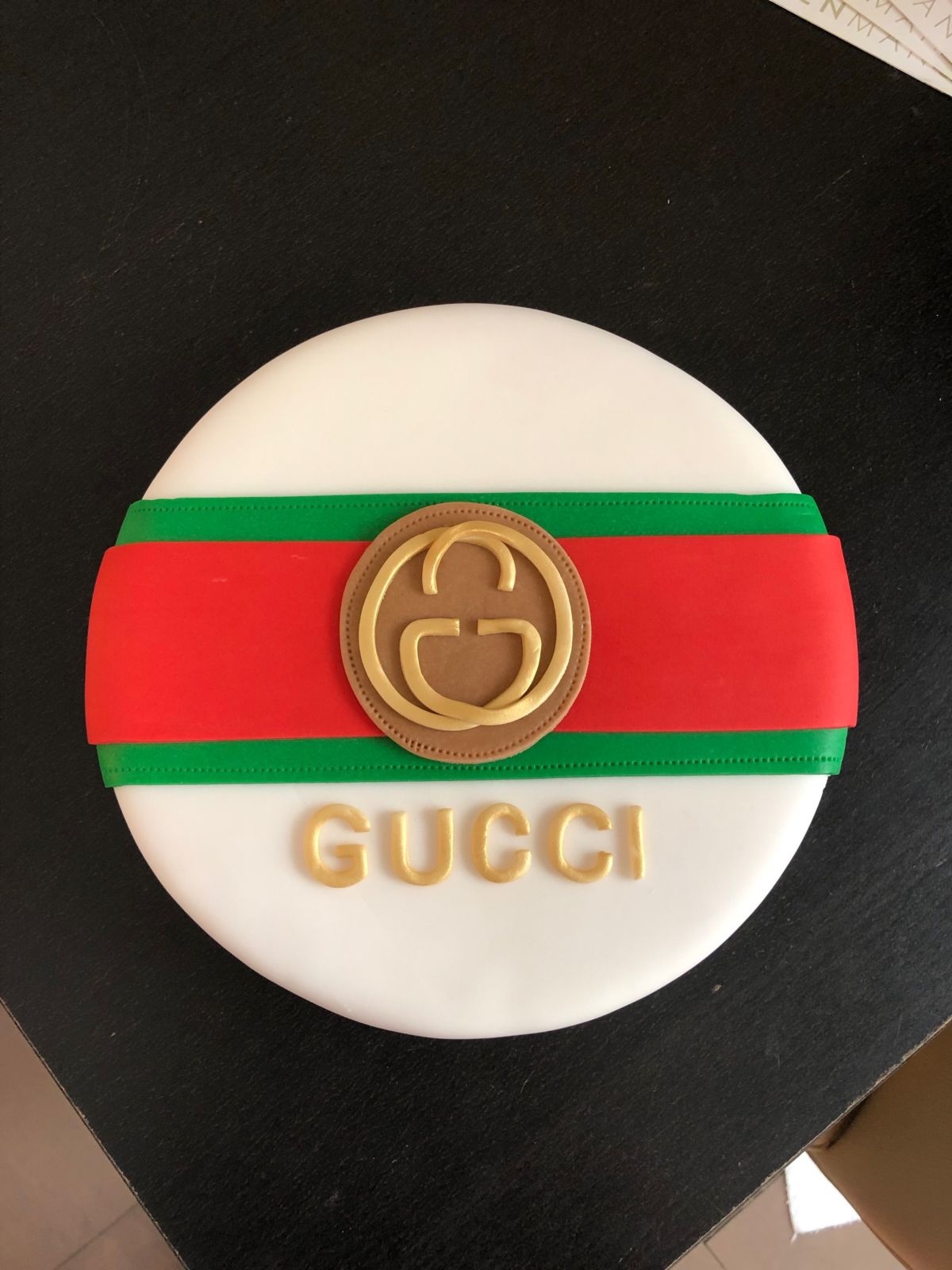 Round cake with white fondant, red and green Gucci brand stripes, gold circular seal and lettering